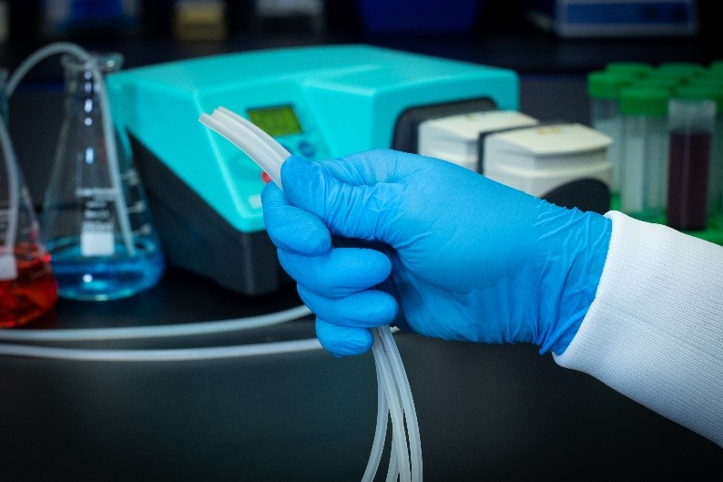 Gloved hand holding flexible tubing in a laboratory setting, with a peristaltic pump and lab equipment, including flasks and test tubes, in the background.